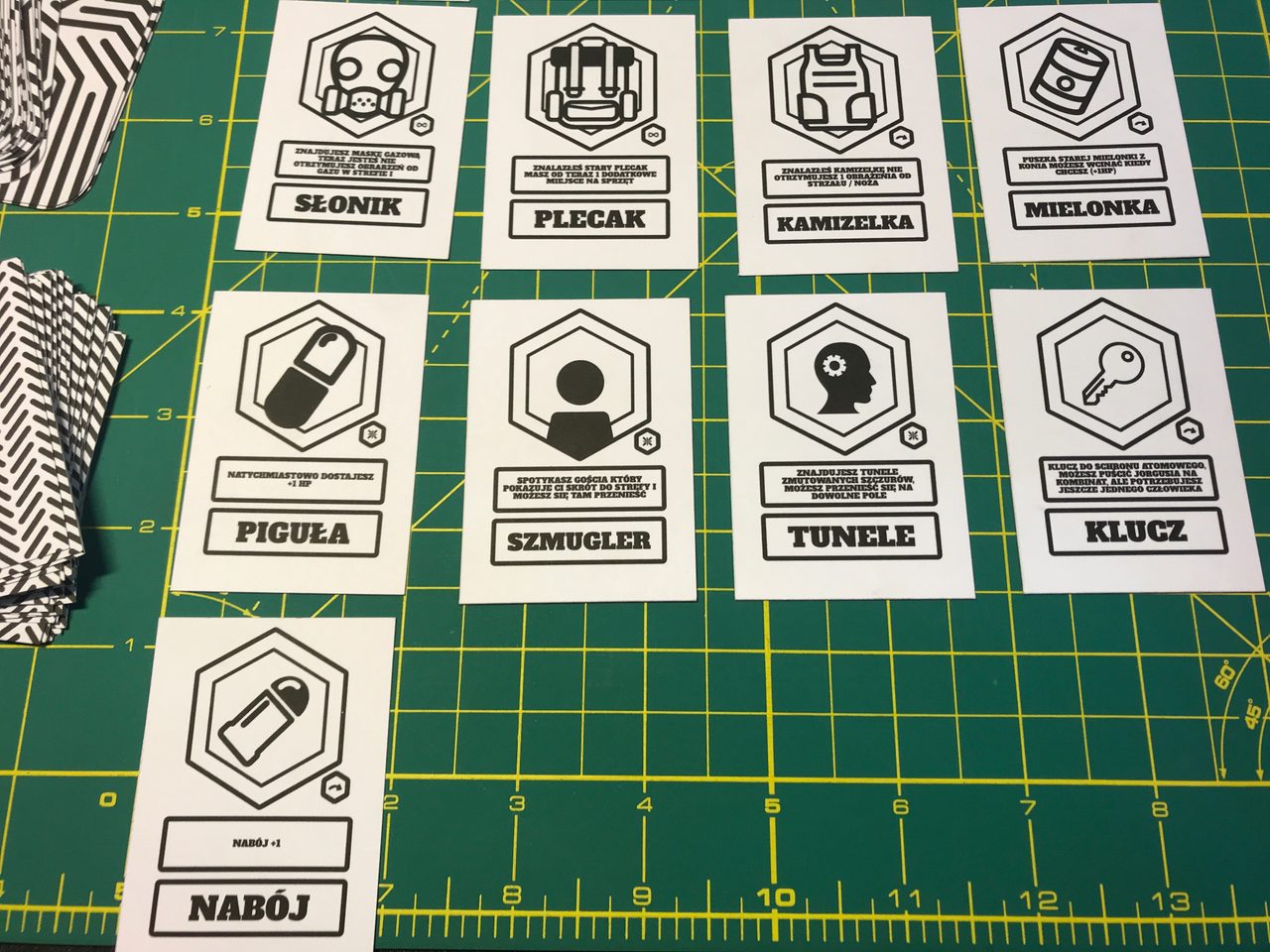 Black-and-white prototype cards for equipment and events arranged on a green cutting mat.