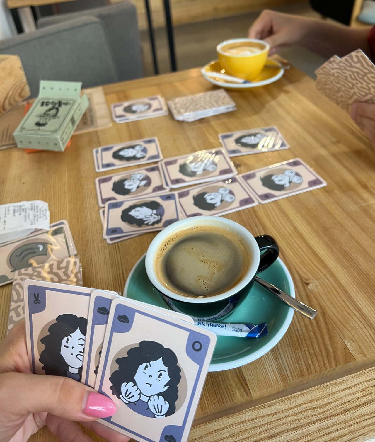 Two people playing ToeShamBo at a cafe, with illustrated cards spread across the table between their coffee cups.