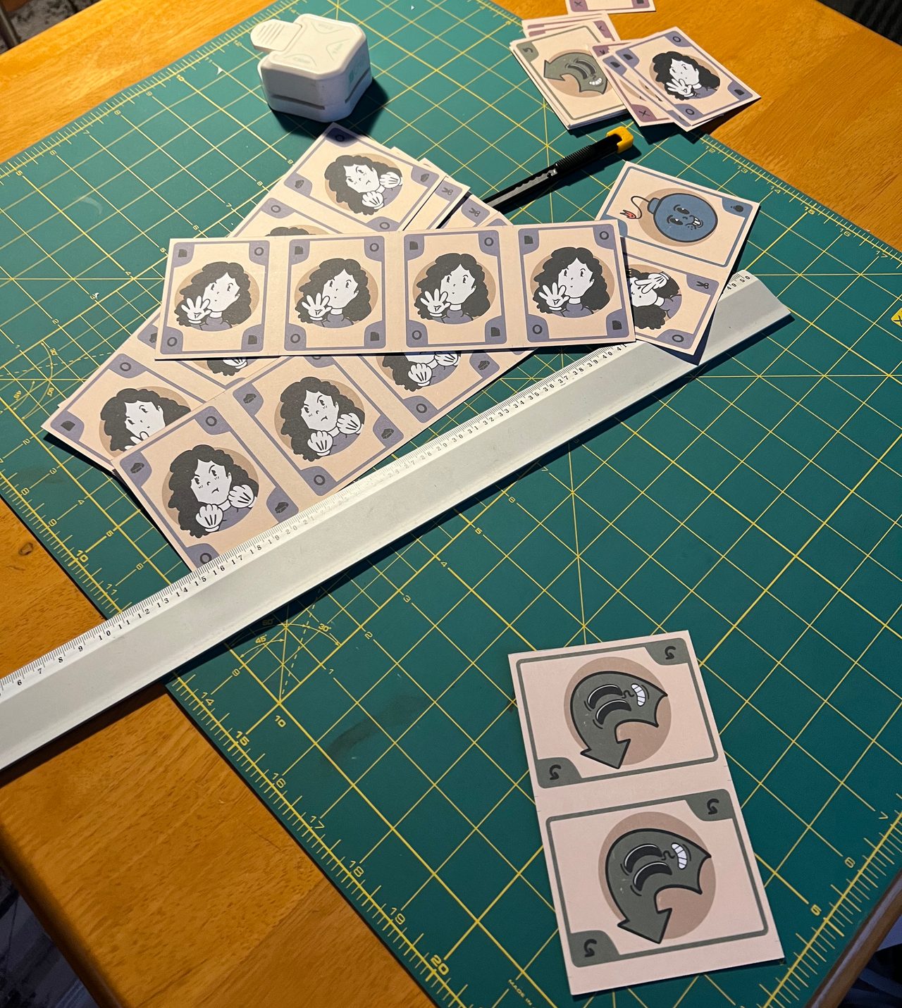 Printed ToeShamBo cards being cut on a green cutting mat with a ruler and craft knife.